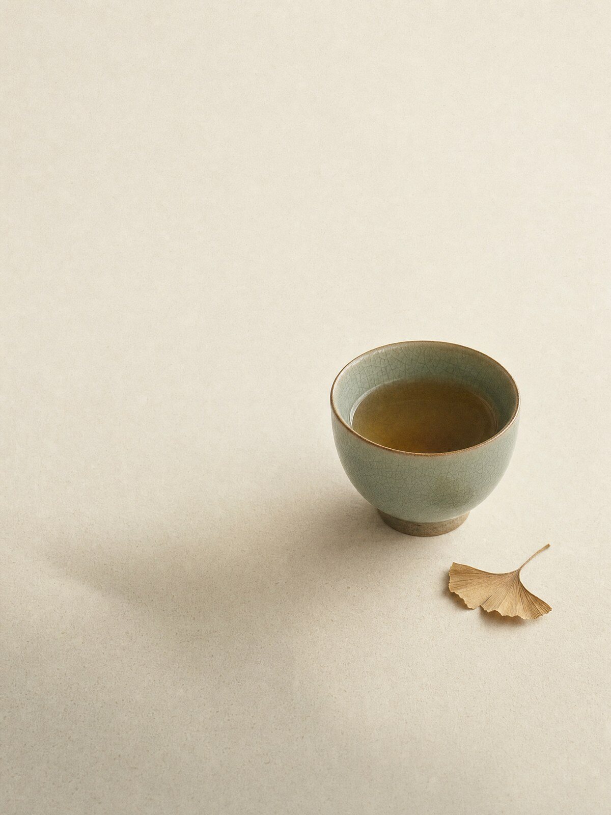 A celadon cup of tea and a single ginkgo leaf on a soft cream surface
